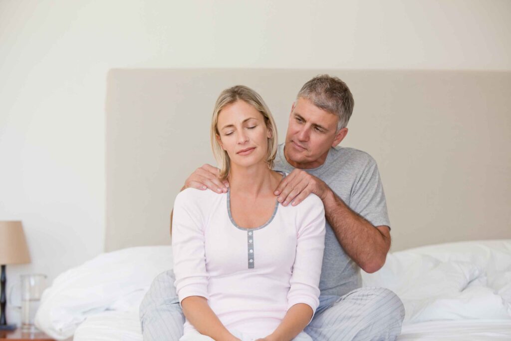 A middle-aged man gently giving a shoulder massage to a relaxed woman sitting on a bed, showing intimacy and trust in a bedroom setting