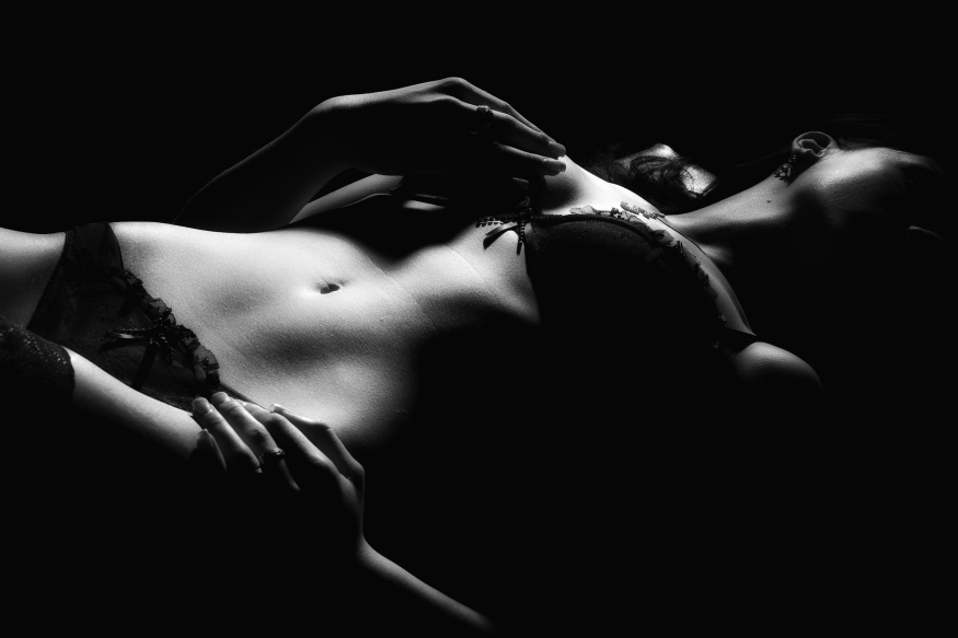 Black and white close-up of a woman lying down in sensual lingerie, with her hands near her chest and stomach, bathed in dramatic light and shadow.